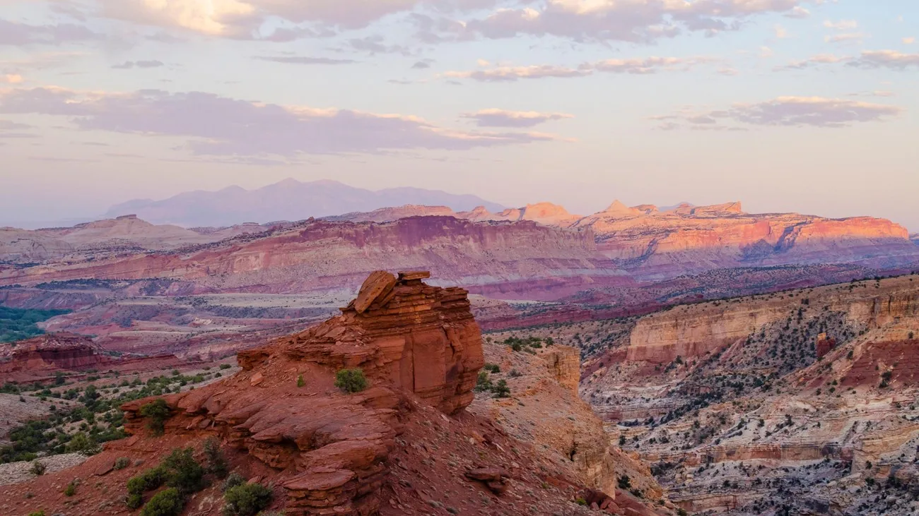 Sunset lights layered red and cream cliffs at Capitol Reef, with a sandstone outcrop in the foreground and distant mountains beneath pastel clouds.